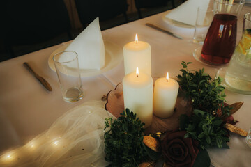 Obraz premium Table setting with three lit candles surrounded by greenery and a decorative rose, elegant dining atmosphere with glassware and cutlery visible in the background