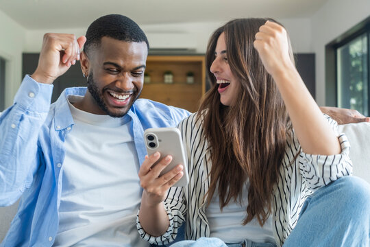 Happy diverse couple cheering and raising fists in joy, celebrating great news, achievement, or a win while looking at a smartphone screen together on a couch at home
