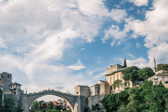 Architectural framing of the historic Stari Most stone bridge and surrounding heritage buildings set against a dynamic cloudscape in Mostar, Bosnia.