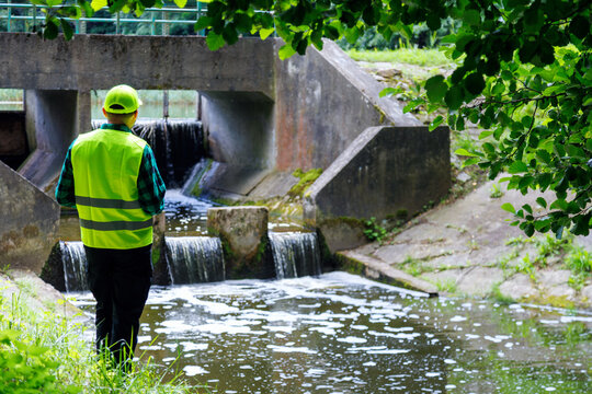 Back view of a hydraulic engineer or environmental inspector in a high-visibility safety vest checking a concrete dam spillway and water flow. Environmental supervisor inspecting water quality.