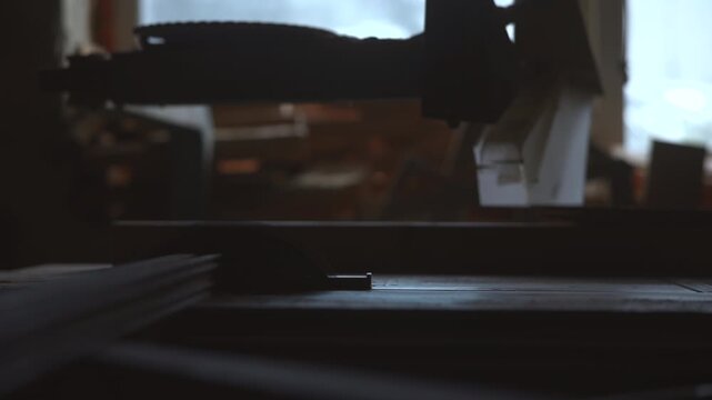 Low-angle workshop view of suspended blade guard above stacked wood with moody daylight, worn textures, and industrial safety details inside dim workspace, with workshop, blade, guard, wood detail