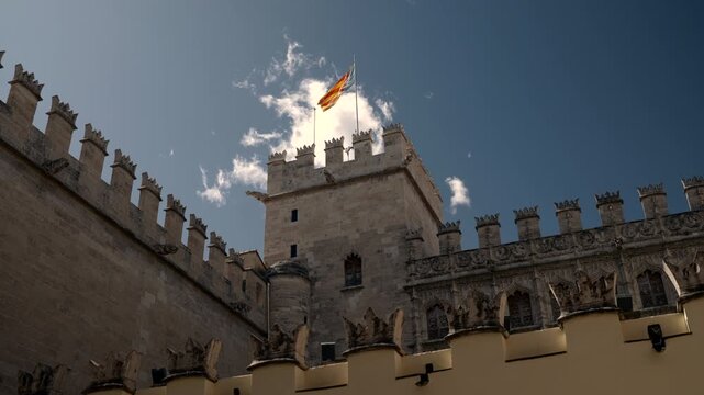 Low angle rooftop tower with unfurled flags above crenellated historic stone parapet, carved cornices, weathered limestone textures and clear blue sky on sunlit heritage landmark