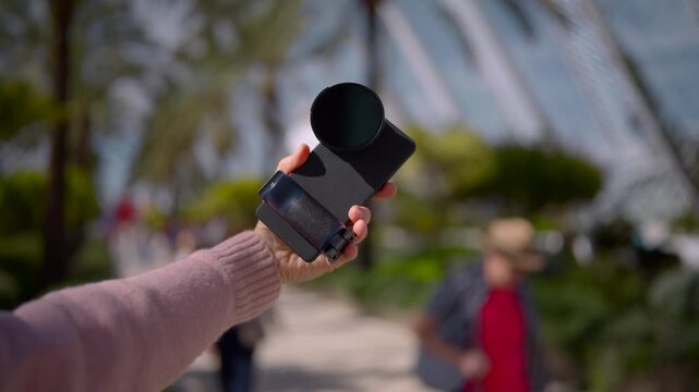 Hand presenting smartphone fitted with compact stabilizing grip and circular filter in sunlit park walkway during calm outdoor demonstration with soft green background
