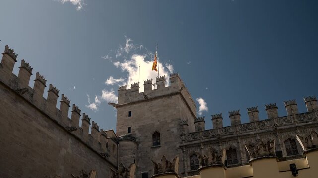 Low angle view of weathered stone fortress tower with fluttering flag, crenellated parapet, carved details, and bright blue sky with drifting clouds, with flag, tower, parapet, crenellation detail