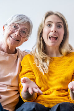 Grandmother and young woman looking directly at camera with wide eyes and open mouths, reacting to something surprising with joyful expressions and shared excitement