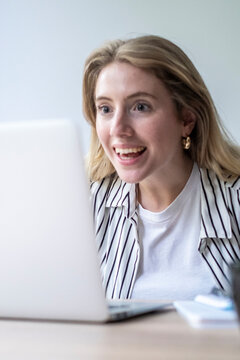 Joyful young woman engaging in a video call on her laptop, expressing surprise and excitement while receiving good news or celebrating success during remote work or online communication