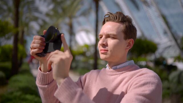 Young man adjusting compact camera outdoors in sunny urban park with focused expression, fine hand movements and careful framing beside modern architecture, with man, person, camera, photography