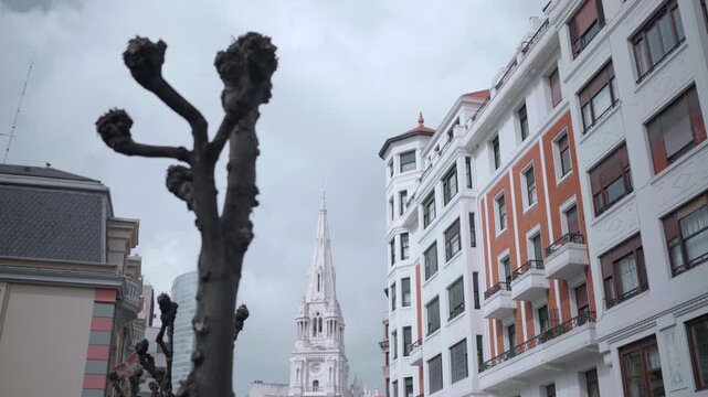 Low angle city street with pollarded trees, ornate brick facades, slate roofs, tall church spire, and muted overcast light creating strong vertical urban composition
