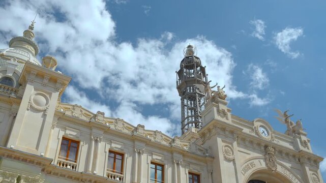 Low angle view of ornate historic building rooftop with cream facade, decorative stonework, openwork metal tower, gilded details, and drifting clouds over blue sky