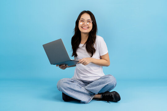 Young woman happily working on a laptop while sitting cross-legged on a blue background
