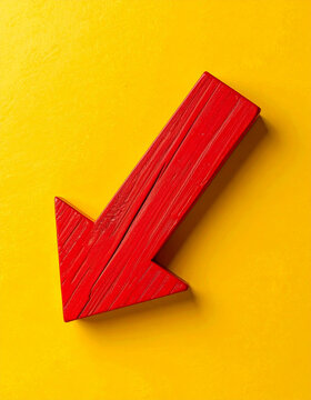 Bold red wooden arrow pointing downward on bright yellow background, symbol of business decline, falling market prices and economic downturn. Close-up.