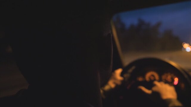 Close up of young guy holds hands on steering wheel and drives a modern automobile at evening. Male driver moves in his SUV car on empty road. Man rides in auto on highway at night time. Slow motion