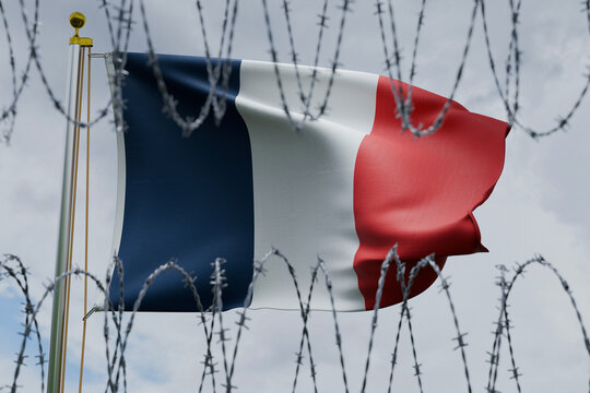 French flag waving on a flagpole, viewed behind sharp coiled barbed wire against a cloudy sky. Conceptual image representing European immigration policy, French border security, and migration control.