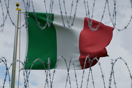 Italian flag waving on a flagpole, behind sharp coiled barbed wire on a cloudy sky. Conceptual image representing European immigration policy, Italian border security, and migration control.