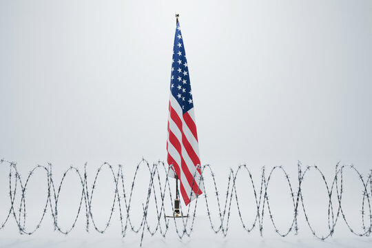 American flag on a stand behind a row of coiled barbed wire against a white studio background. Conceptual metaphor for US immigration policy, illegal migration, and border security.