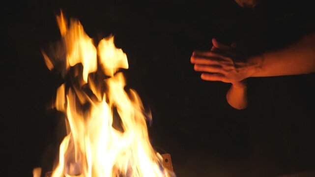 Low view of young tourist rubs his hands in front of an open bonfire from mangal at night. Man warms his arms by a fire from metal brazier against the black background. Concept of warmth and tranquil