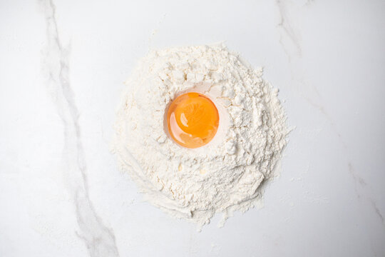 A cracked egg with a bright orange yolk sits in the center of a mound of white flour, viewed from above on a marble surface