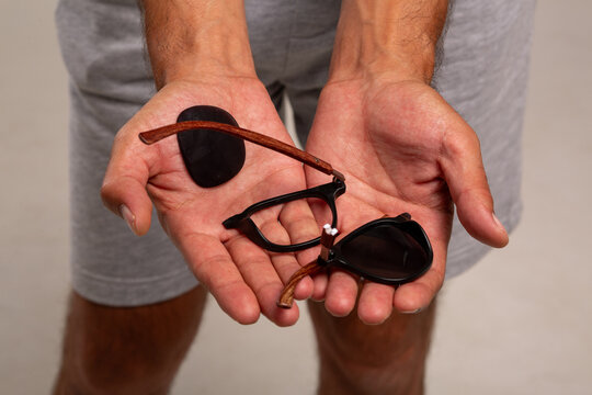 A young guy in a colored T-shirt and gray shorts holds broken glasses in his hands after unsuccessfully transporting luggage at the airport. The glasses are made of low-quality material