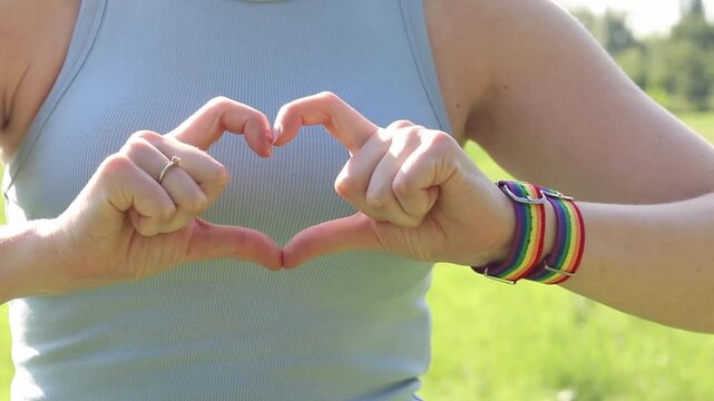 Female hands show heart gesture on blurred natural background. Woman with rainbow LGBTQ+ community bracelet on wrist. Girl makes heart shape with fingers, close-up. Love and tolerance concept