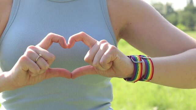 Female hands show heart gesture on blurred natural background. Woman with rainbow LGBTQ+ community bracelet on wrist. Girl makes heart shape with fingers, close-up. Love and tolerance concept