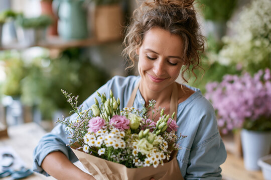 Smiling florist holds a vibrant bouquet wrapped in brown paper. Her curly hair frames her face as she looks down. Soft lighting highlights the delicate pink roses and white daisies