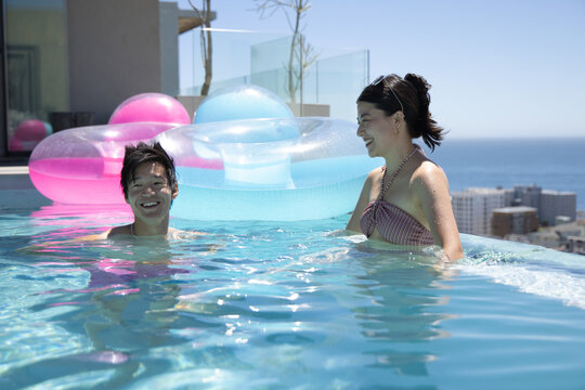 Asian couple laughing and swimming in rooftop infinity pool near floats, woman in striped bikini