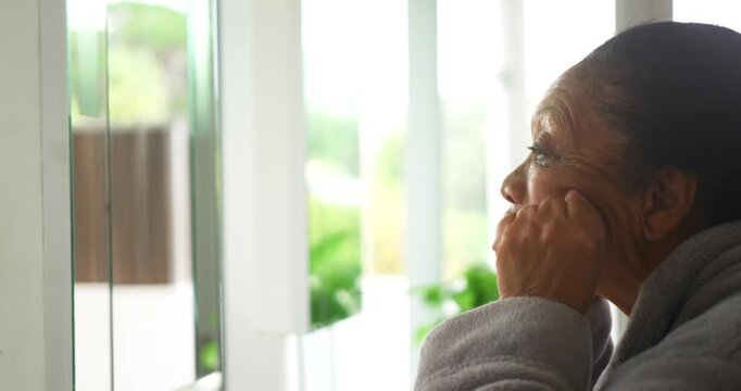 Senior African American woman sitting at blinds at home wearing gray robe seeing yard resting chin