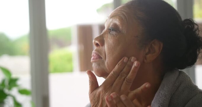 Senior African American woman gray robe pressing skincare around face by glass door sunlight urging