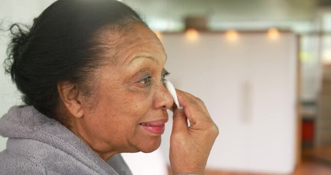 Senior African American woman picking pad, cleansing skin in kitchen wearing grey robe, copy space