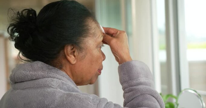 Senior African American woman in gray robe standing at sink by window raising pad removing makeup