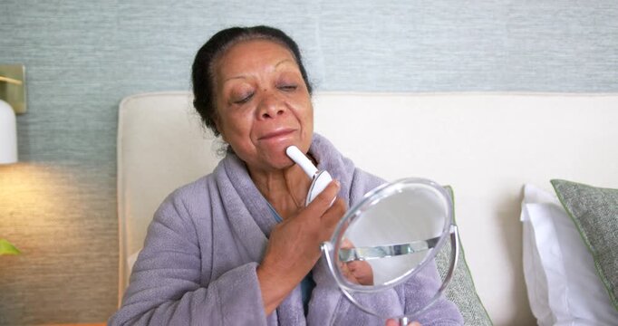 Senior African American woman in grey robe starting skincare on bed, holding mirror, gliding device