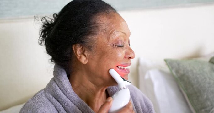 Senior African American woman on bed in gray bathrobe activating T-shaped device treating jawline