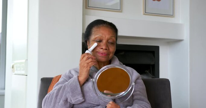 Senior African American woman checking mirror massaging eye with skincare device at home for skin