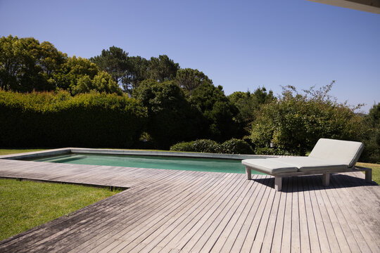 Rectangular pool holding muted blue water on wide wood deck, chaise near grass, hedge casting shade