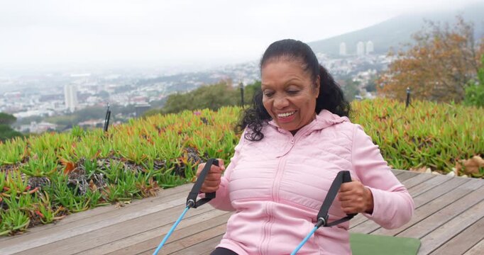 Senior African American woman pink-jacket holding blue band rowing seated on mat on deck exercising