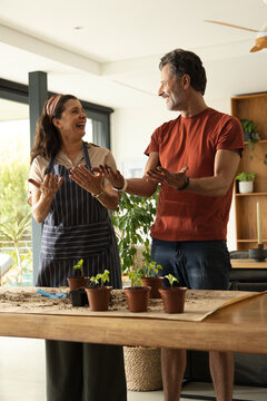 Couple potting seedlings at kitchen table with clay pots, blue trowel, smiling, wearing apron