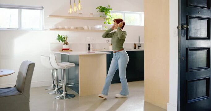 Woman ending call, putting on headphones, wearing green top, dancing in kitchen to phone music