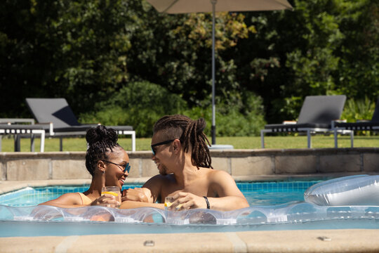 African American couple leaning on float poolside in swimsuit and shirtless holding yellow drinks