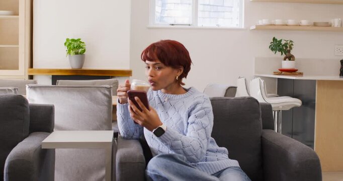 African American woman sitting in chair holding phone, cup, checking texts after alert, in sweater