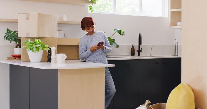 African American woman leaning on island in kitchen, scrolling phone while alert pinging near boxes