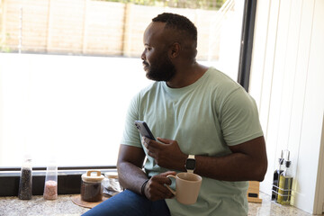 Mid-adult African man sitting on granite countertop by bright window, holding mug and smartphone © wavebreak3