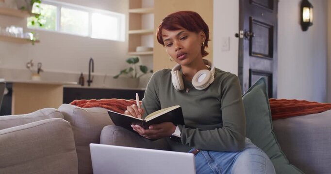 Adult African woman sitting on couch writing in notebook glancing at laptop white headset for study