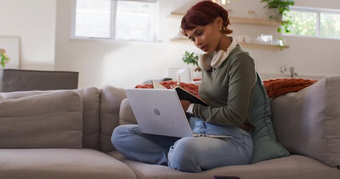 African American woman placing laptop on lap, reading, writing in notebook on gray sofa, copy space