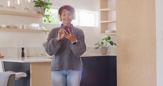 African American woman tapping smartphone, hearing music through headphones and dancing in kitchen