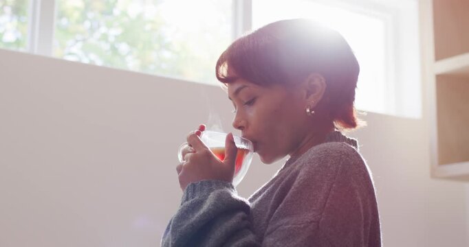 African American woman wearing knit sweater noticing steam, cupping glass cup by window, inhaling