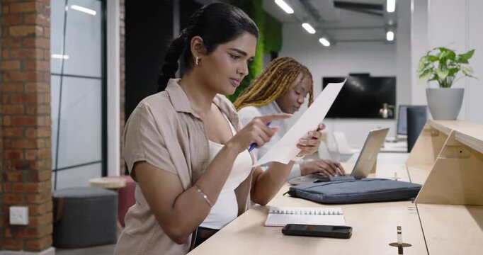 Indian and African women at desk, Indian opening notes marking chart while African typing on laptop