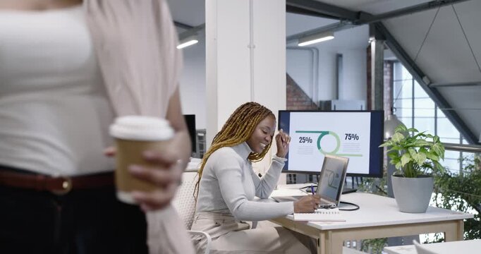 Seated African woman responding to colleague at desk, pointing 25% or 75% screen, using laptop notes
