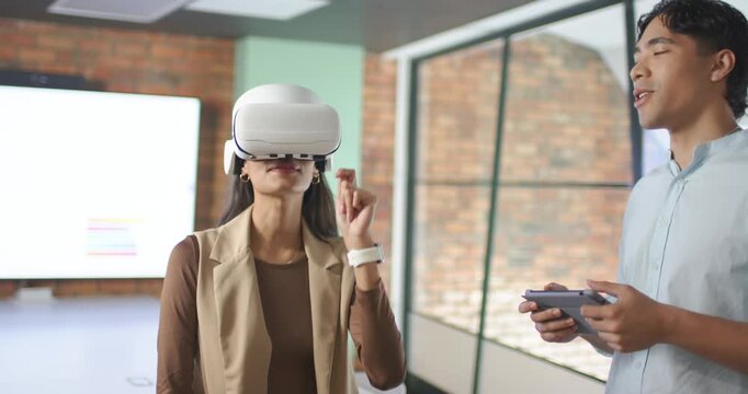Diverse coworkers, man guiding, woman testing VR with white headset as he holds tablet in office