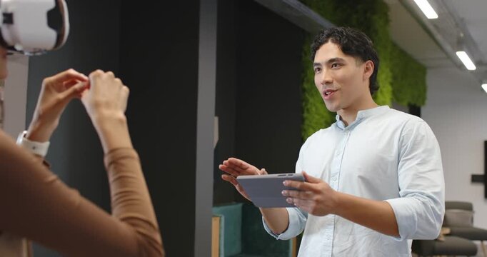 Diverse team in office guiding VR headset, man tapping tablet, woman raising gestures by moss wall