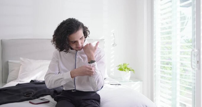 Man adjusting white shirt cuff and fastening black wristwatch on bed preparing for work